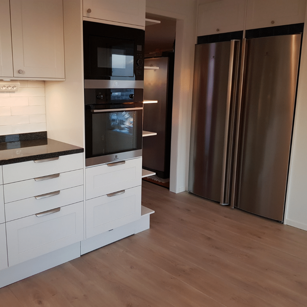 Vitt kök med dubbla kylskåp i rostfritt stål och inbyggda ugnar, en modern köksrenovering som fokuserar på funktion och förvaring.