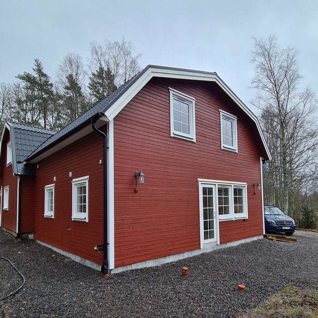 Ett stort, falurött trähus med vita fönsterkarmar och svarta takpannor. Huset har ett brutet tak och står på en grusad uppfart med en skog i bakgrunden.