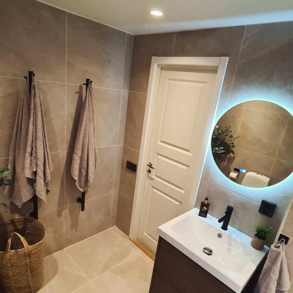 Modernt badrum med beige kakel, handdukshängare och rund spegel med LED-belysning – byggföretag i Norrköping