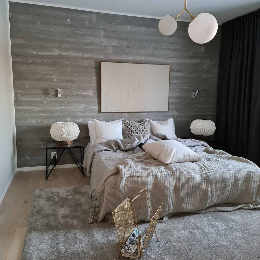 Ett sovrum med en fondvägg av grå, liggande träpanel. En dubbelsäng är bäddad med ljusa sängkläder och över sängen hänger en stor, vit tavla. En modern taklampa med vita klot och mässingsdetaljer hänger från taket.