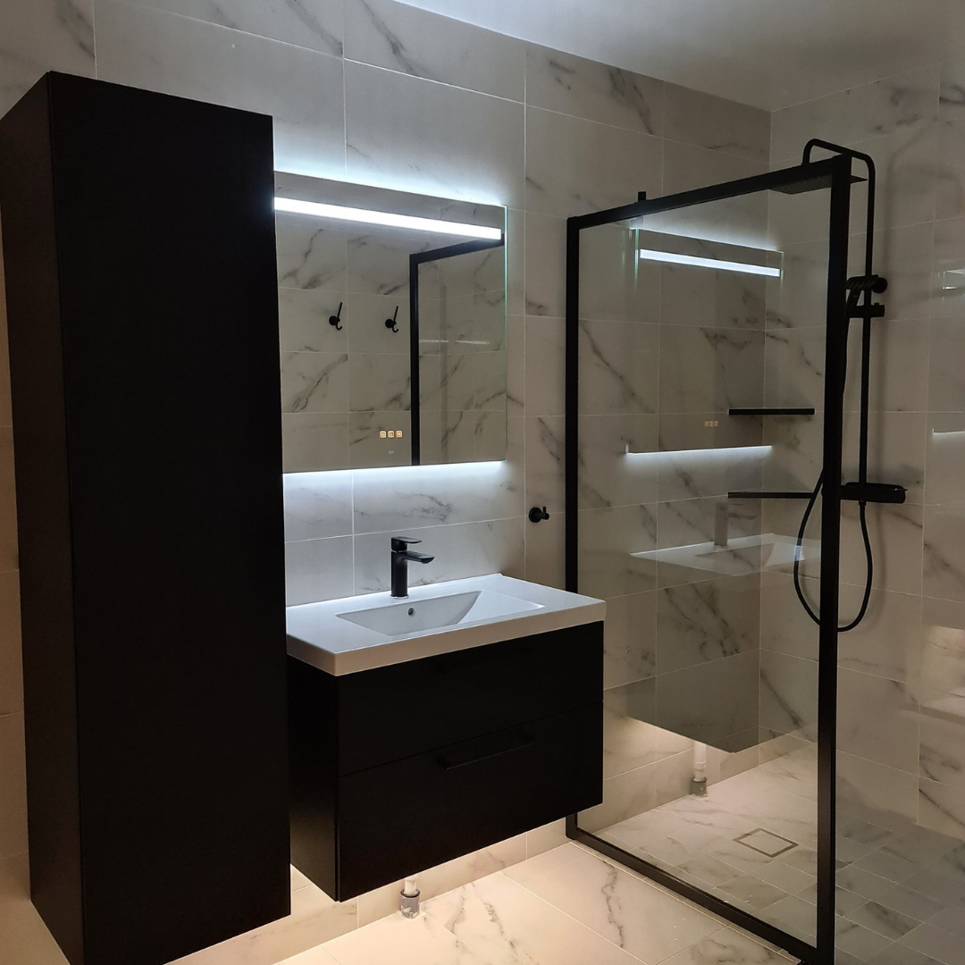 Ett badrum med vit marmorklinker, svarta badrumsmöbler och en spegel med integrerad LED-belysning. Duschväggen har svarta profiler och en svart takdusch.