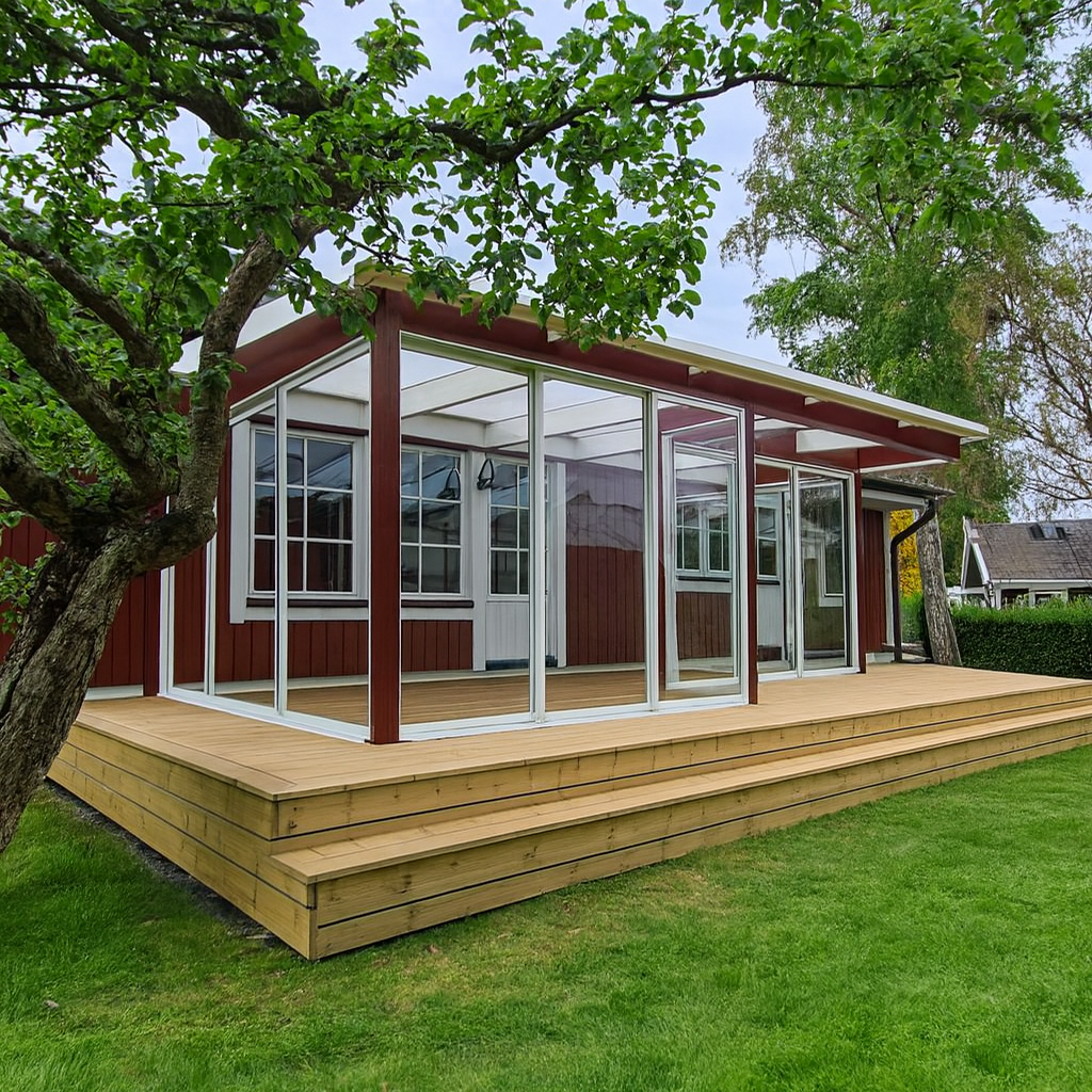 A red wooden house with a newly constructed, multi-tiered wooden deck and an attached glass conservatory with white frames. The deck leads down to a vibrant green lawn, surrounded by trees, showcasing a harmonious blend of traditional architecture and modern outdoor living.