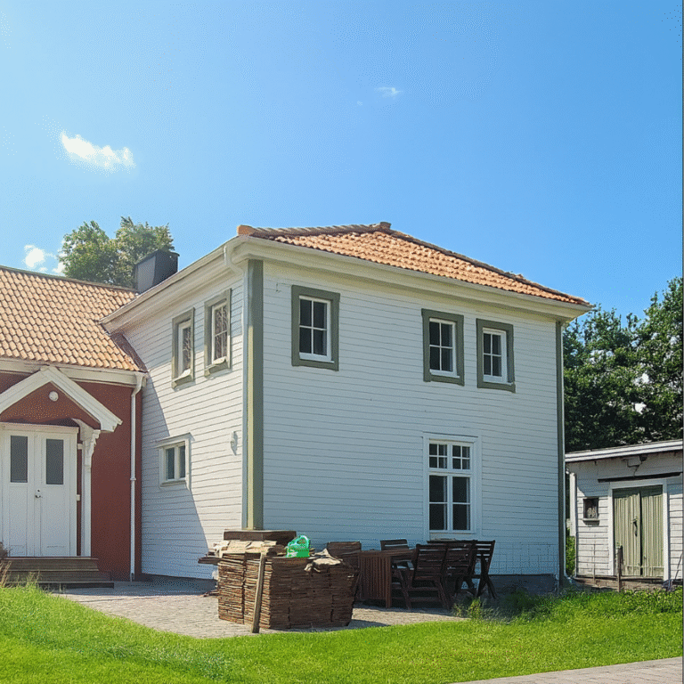 A white wooden house with green window trim and a red tiled roof, connected to a red section of the house. A wooden outdoor dining set and stacked wooden planks are visible on a paved area, surrounded by green lawn under a clear blue sky, indicating a renovated or extended property.