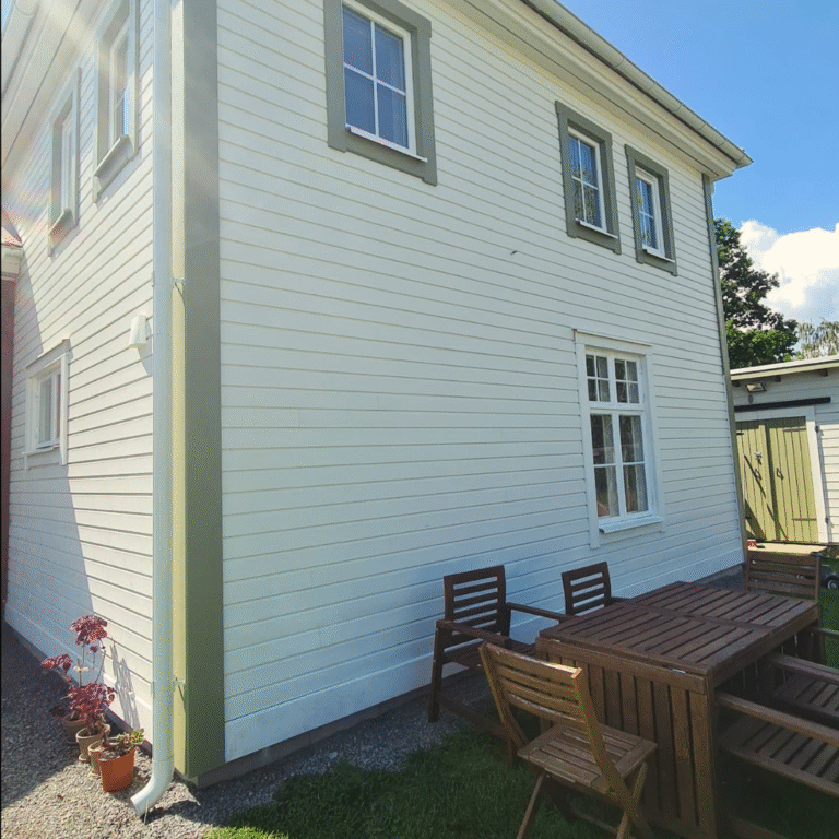The side of a white wooden house with green trim around the windows and a green downspout. A wooden outdoor dining table and chairs are set on a grassy patch next to the house, suggesting a pleasant outdoor living area.