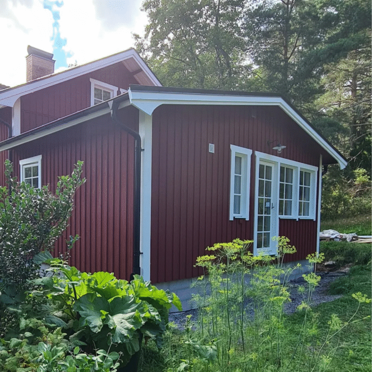 A charming red wooden house with white window frames and trim, featuring a newly added extension. The house is surrounded by vibrant green plants and trees under a bright sky, showcasing traditional Scandinavian architecture and home improvements.