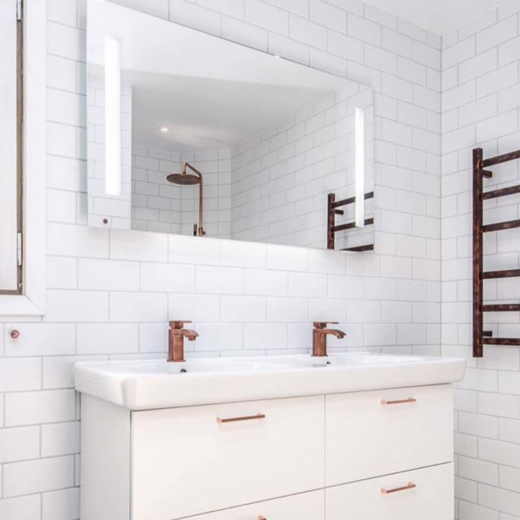 A modern bathroom with white subway tiled walls, featuring a double vanity with two white sinks and copper-colored faucets. Above the vanity is a large rectangular mirror with integrated lighting, reflecting a shower head. A copper-colored heated towel rail is mounted on the right wall, showcasing a cohesive and stylish bathroom design.