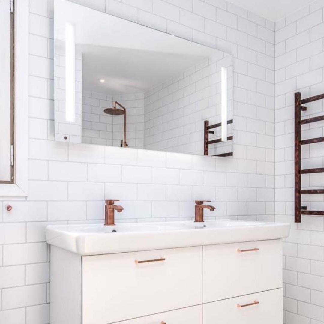 A modern bathroom with white subway tiled walls, featuring a double vanity with two white sinks and copper-colored faucets. Above the vanity is a large rectangular mirror with integrated lighting, reflecting a shower head. A copper-colored heated towel rail is mounted on the right wall, showcasing a cohesive and stylish bathroom design.
