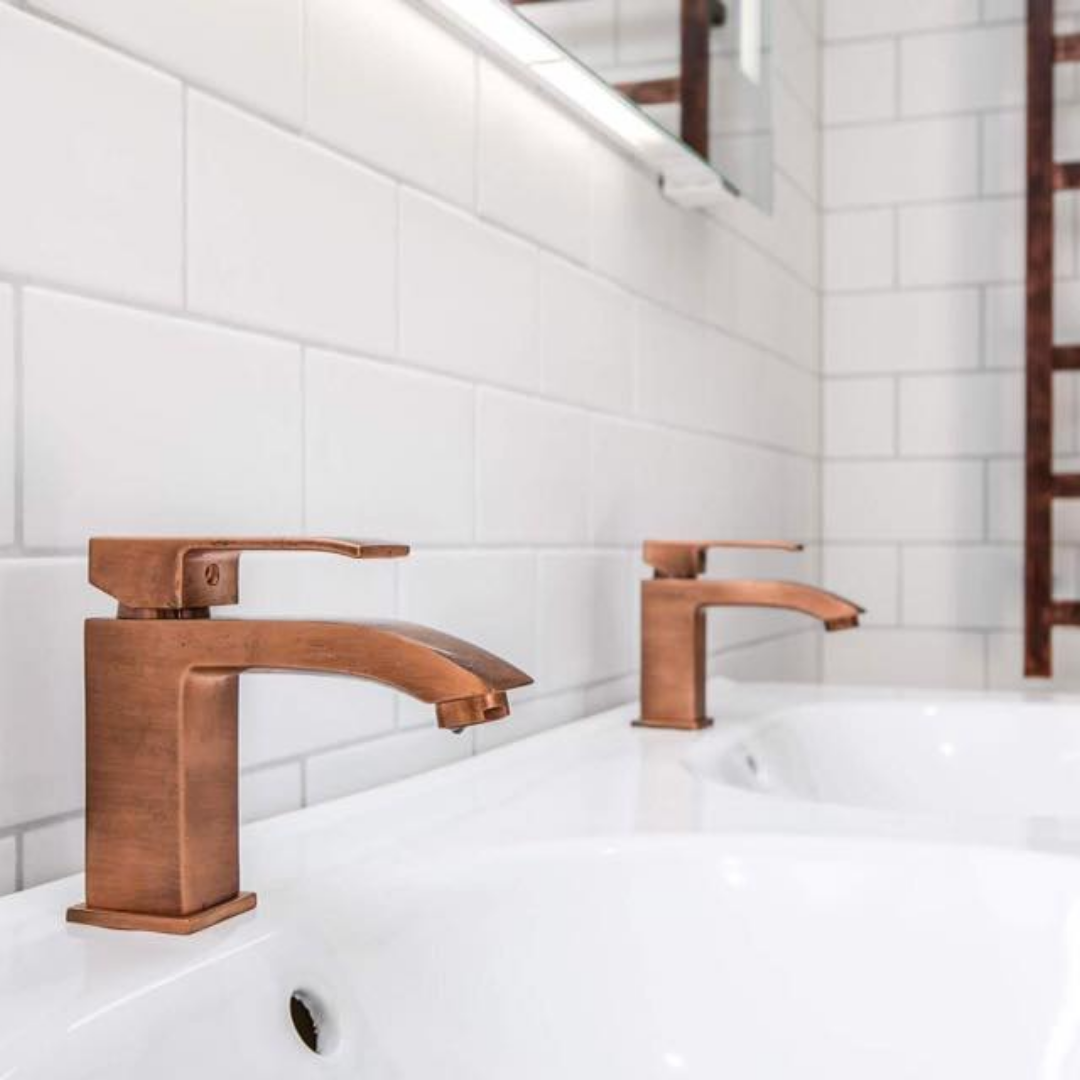 A close-up of two modern, square-shaped copper-colored faucets over white ceramic sinks, set against a backdrop of white subway tiles. A heated towel rail is partially visible in the background, emphasizing elegant bathroom fixtures and design details.