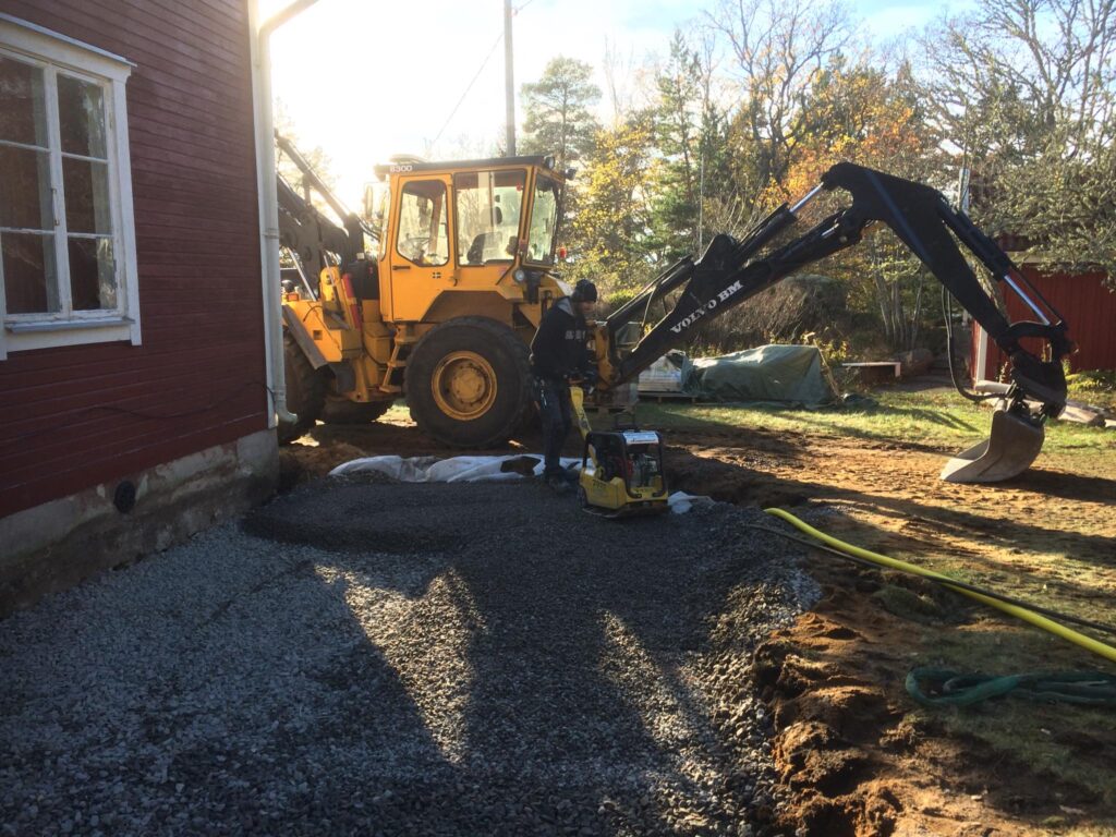 A construction worker operating a plate compactor on a layer of gravel next to a red wooden house. A yellow Volvo excavator is parked in the background, and the scene is bathed in warm sunlight, depicting ground preparation for a new construction project or landscaping.