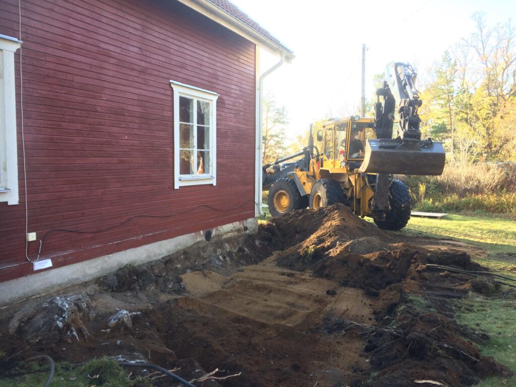A red wooden house with white trim, next to an excavation site where a yellow Volvo excavator is digging. Piles of dirt are visible, indicating ground work for a new foundation or extension, set against a backdrop of autumn trees.