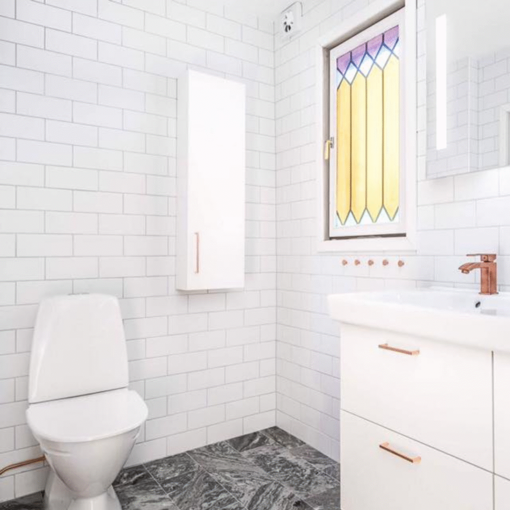 A contemporary bathroom featuring white subway tiles on the walls and dark patterned floor tiles. A white toilet is on the left, and a white vanity with a sink and copper-colored faucets is on the right. A window with colorful stained-glass film and a mirror with integrated lighting add unique touches.