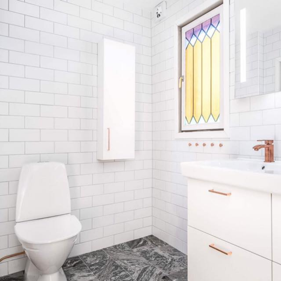 A contemporary bathroom featuring white subway tiles on the walls and dark patterned floor tiles. A white toilet is on the left, and a white vanity with a sink and copper-colored faucets is on the right. A window with colorful stained-glass film and a mirror with integrated lighting add unique touches.