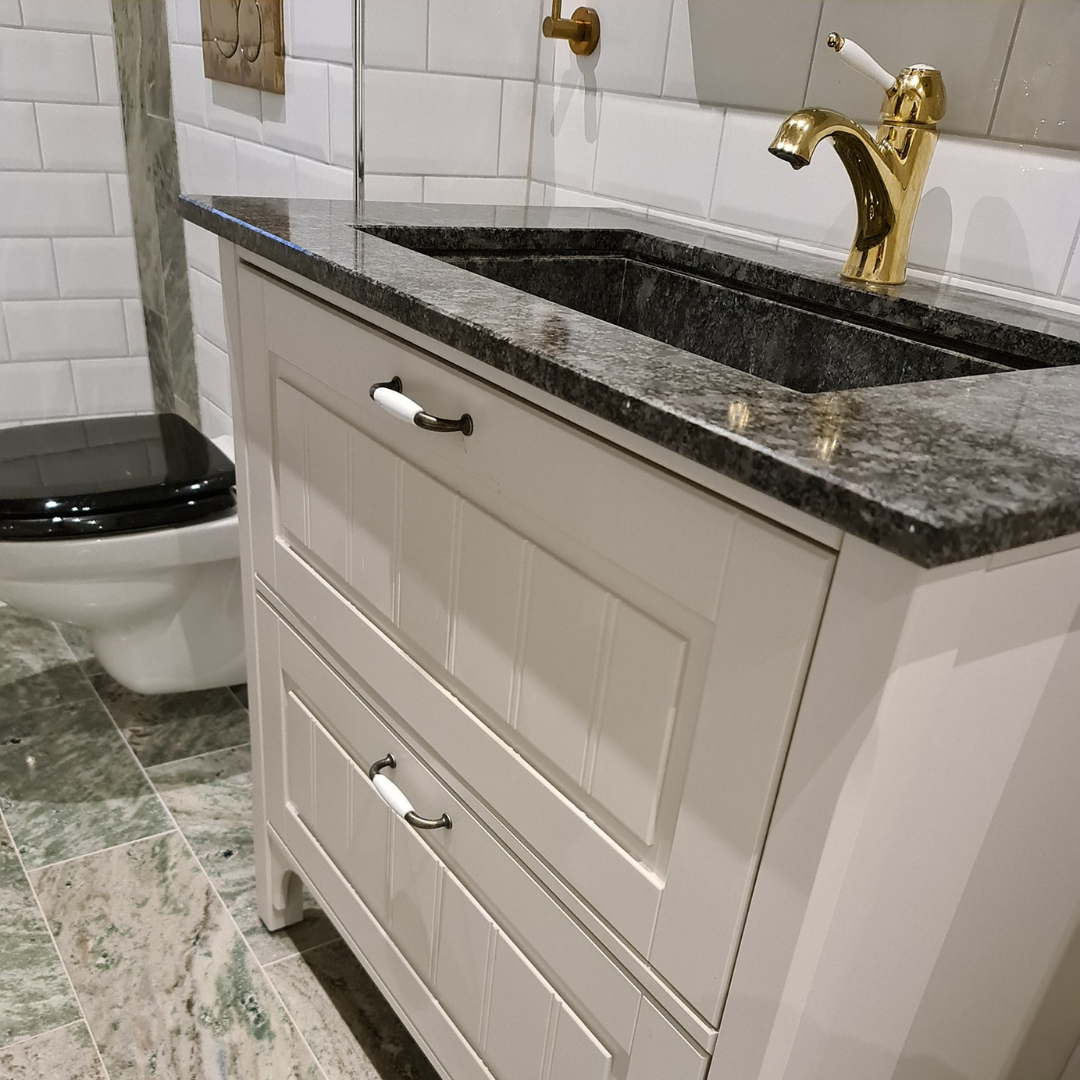 A close-up of a stylish bathroom vanity with a white cabinet, granite countertop, and a black stone sink. A luxurious gold faucet stands out against the white subway tiles and green marble floor, with a black toilet seat visible in the background, highlighting modern bathroom design.