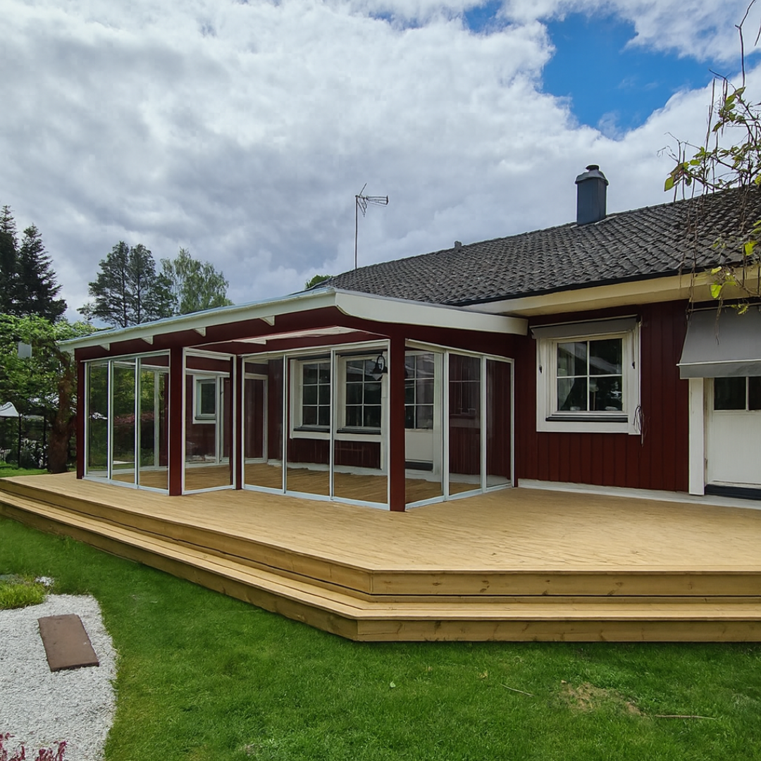 A red house with a newly built, expansive wooden deck and an attached glass conservatory with red framing. The multi-level deck provides ample outdoor living space, surrounded by a lush green lawn and trees under a partly cloudy sky, illustrating a beautiful home renovation and outdoor addition.