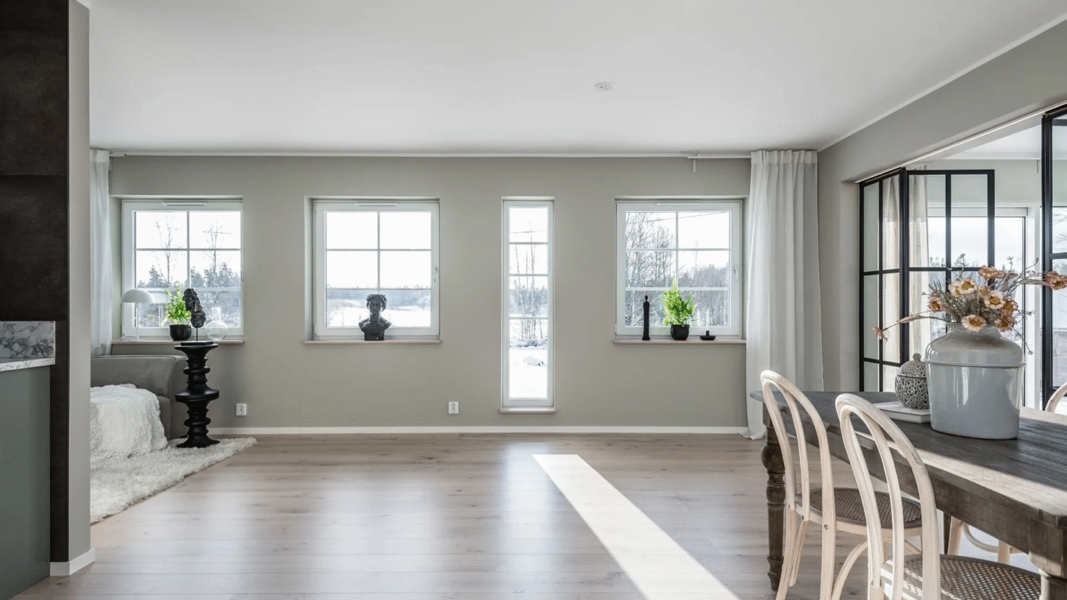 A bright, empty living and dining area with light gray walls and light wooden flooring. Multiple windows offer views of a snowy landscape, and a dark-framed glass partition separates the dining space, highlighting modern interior design and natural light.
