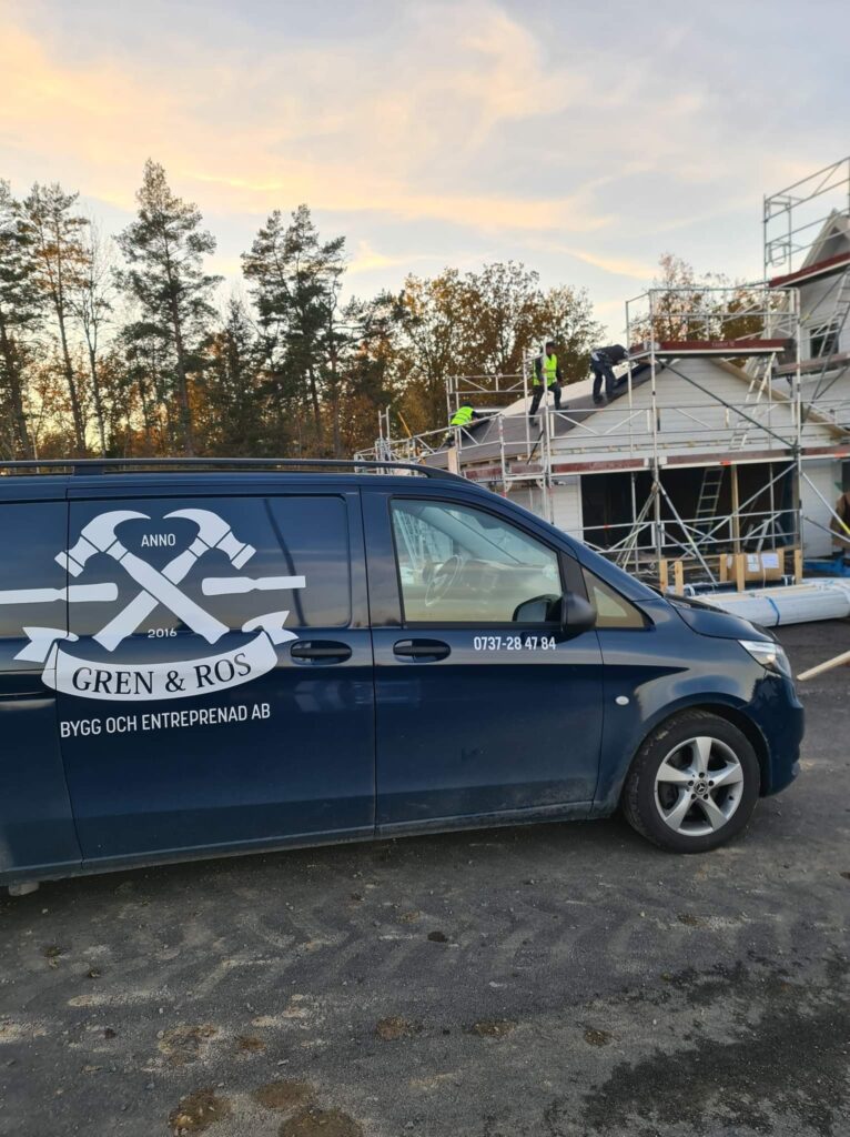 A dark blue Gren & Ros Bygg och Entreprenad AB company van, featuring their logo and contact number, parked at a construction site where workers are actively building a house on scaffolding under a cloudy sky. This image represents professional construction services and building projects.