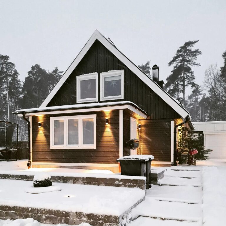 A dark brown house with a prominent gabled roof, covered in snow, with white window frames and trim. Warm lights illuminate the entrance and windows, creating a cozy atmosphere during a snowy evening. Pine trees are visible in the background, highlighting a winter home setting.