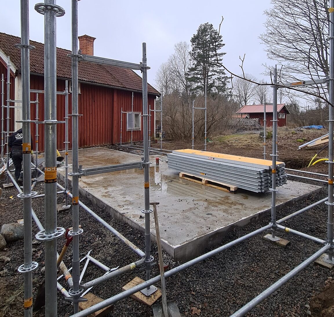 A construction site with metal scaffolding erected around a concrete foundation, next to a traditional red wooden house. Stacks of building materials are on a pallet on the foundation, and bare trees are in the background, illustrating the early stages of a building extension or new construction project.