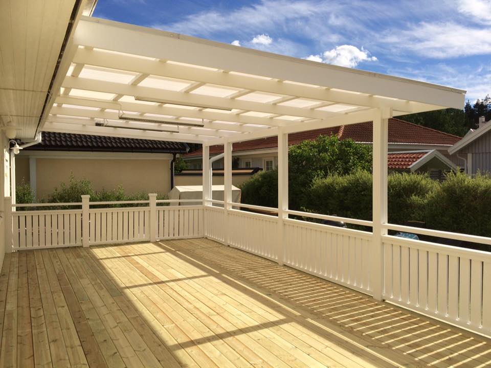 A large, newly constructed wooden deck with white railings and a translucent roof, offering a bright and sheltered outdoor area. The deck overlooks other residential houses and green foliage under a clear blue sky with white clouds, perfect for relaxation and entertaining.