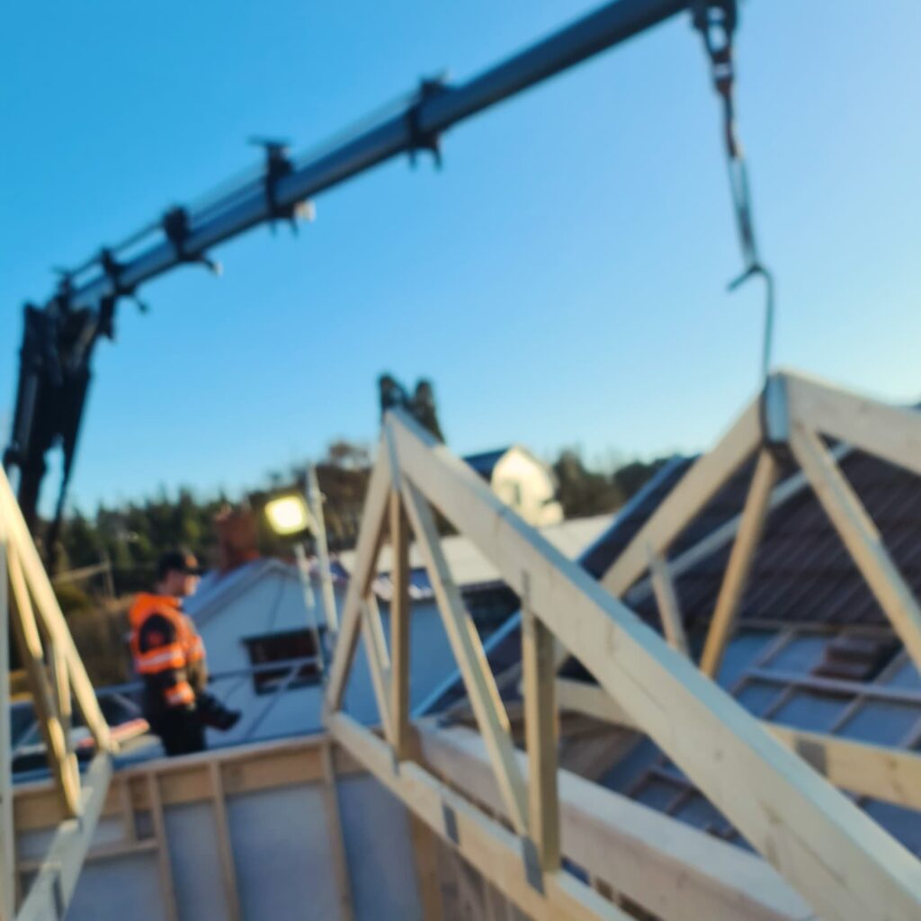 A construction worker in an orange safety vest stands on a building frame as a crane lifts wooden roof trusses into place against a clear blue sky. This image captures the installation of a new roof structure.