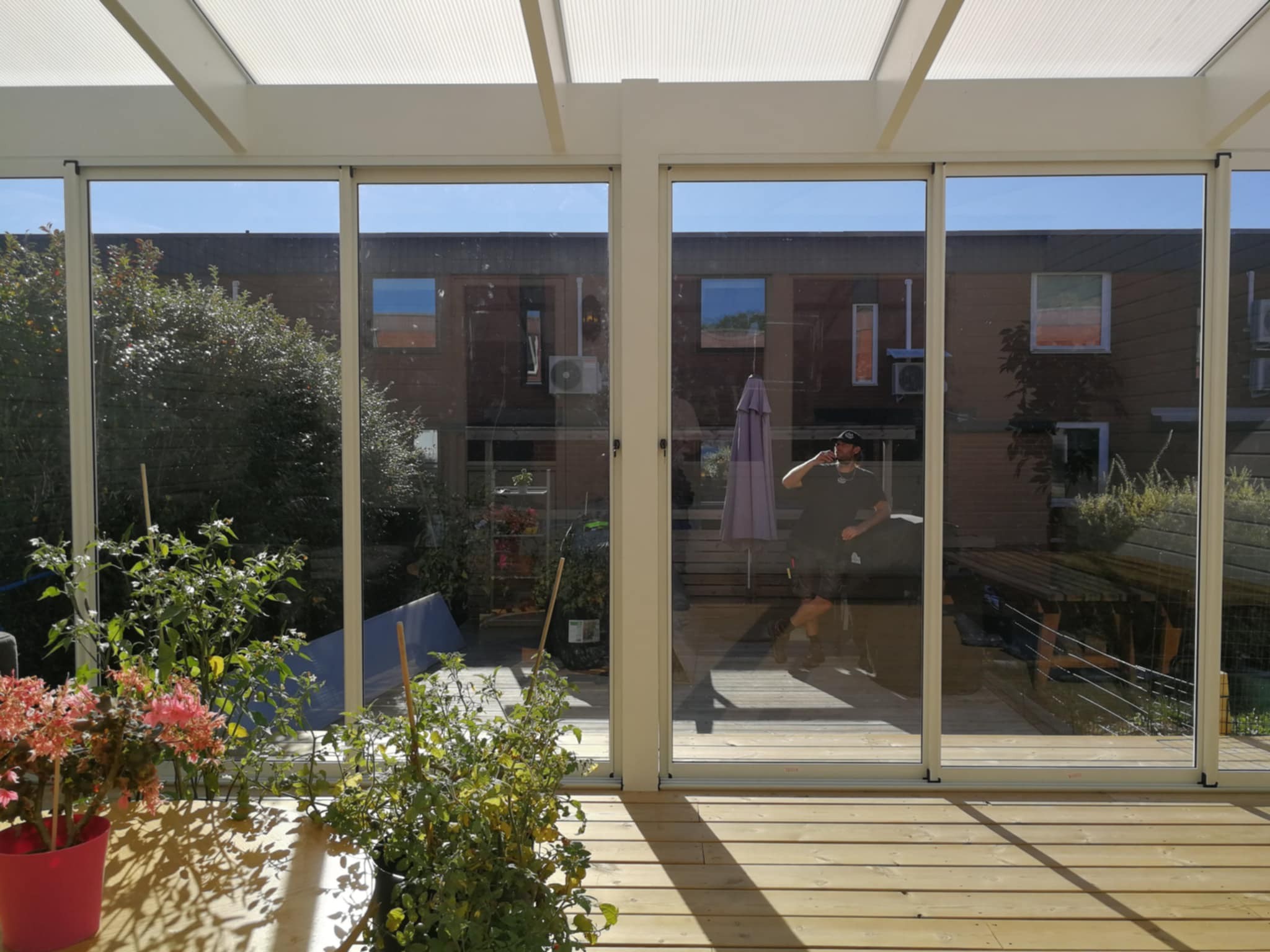 The interior view of a glass-enclosed sunroom or conservatory with a wooden deck floor. Potted plants are visible in the foreground, and through the glass panels, a reflection of a person and the exterior of a house with a garden and outdoor furniture can be seen, creating a bright and airy indoor-outdoor space.