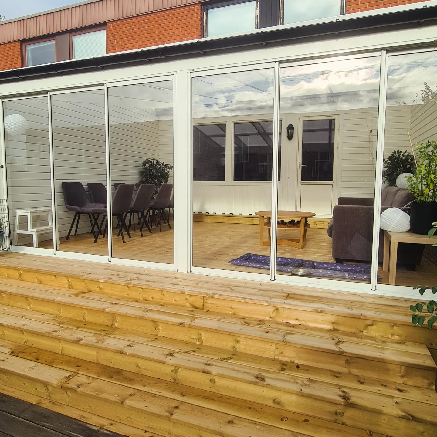 A spacious wooden deck with glass-enclosed sunroom extending from a white house with a brick upper level. The sunroom contains a dining area and lounge furniture, showcasing a bright and versatile outdoor living space.