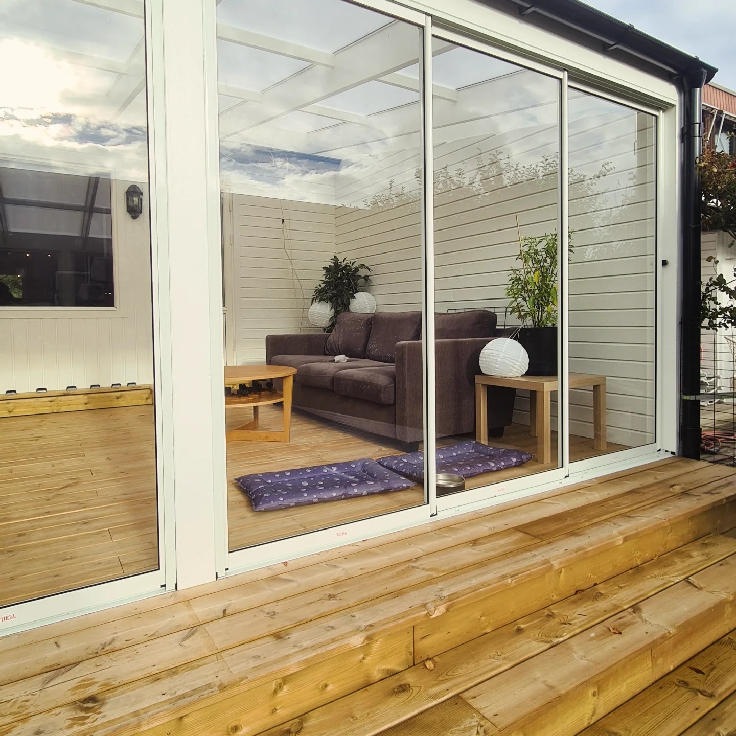 A modern glass conservatory or sunroom with white frames, featuring a comfortable sofa, potted plants, and pet beds on a light wooden floor. The exterior shows a wooden deck leading up to the sunroom, highlighting a cozy and bright indoor-outdoor living space.