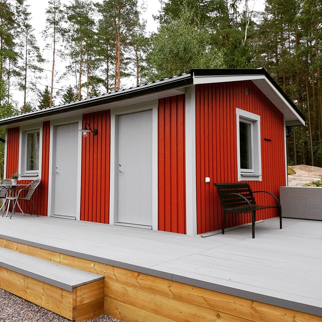 A red wooden cabin with a gray deck, featuring two gray doors and white-framed windows. Outdoor seating is visible on the deck, and the cabin is surrounded by tall pine trees, suggesting a cozy forest retreat or guesthouse.