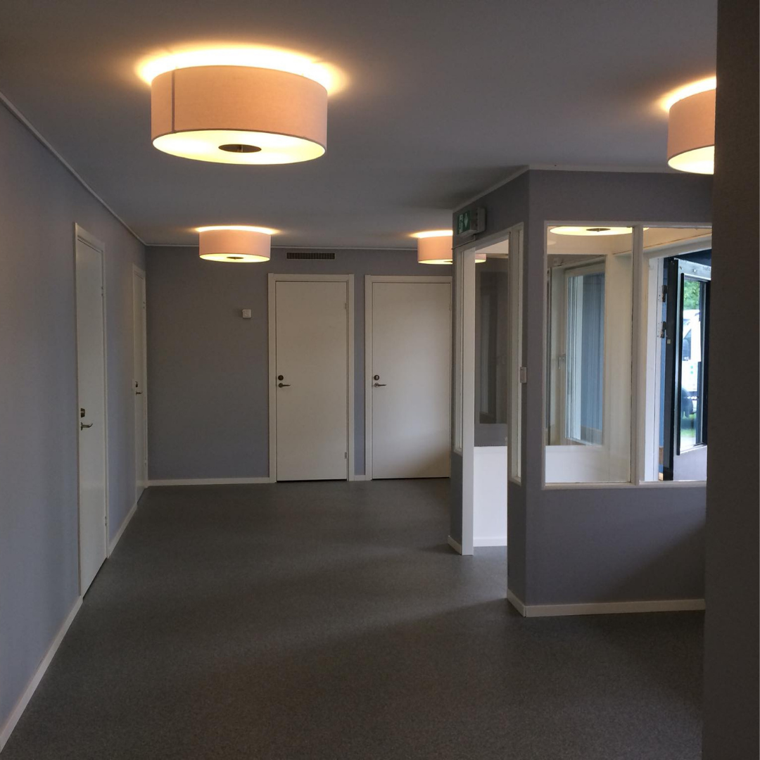 A modern hallway with light gray walls and a gray floor, featuring multiple white doors and contemporary circular ceiling lights. A partial view of an entrance area with glass panels and a radiator is visible on the right, suggesting a newly renovated or commercial interior.