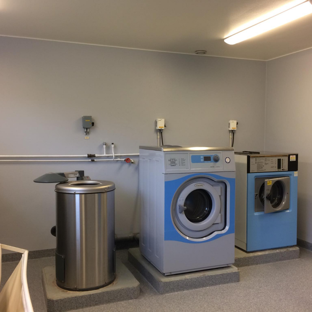 A clean and functional laundry room with light gray walls and a gray speckled floor. It features a large stainless steel bin, a blue and white commercial washing machine, and a blue commercial dryer. Exposed pipes and electrical outlets are visible on the wall, indicating a well-equipped communal or commercial laundry facility.
