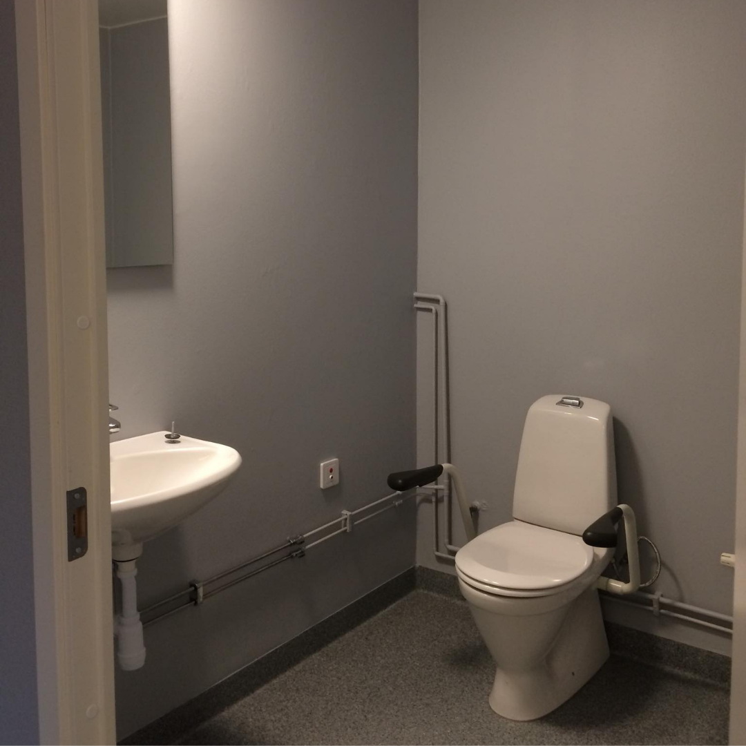 A simple, modern restroom with light gray walls and a gray speckled floor. A white wall-mounted sink with a mirror above it is on the left, and a white toilet with black armrests for accessibility is on the right. Exposed pipes run along the bottom of the walls, suggesting a functional and accessible design.