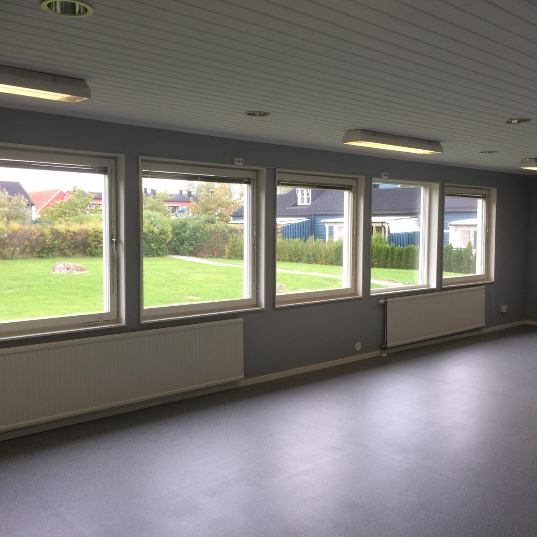 A spacious room with gray walls and a white paneled ceiling, featuring a row of four large windows that offer a view of a green lawn and residential houses outside. White radiators are installed beneath each window, and fluorescent lights are on the ceiling, depicting a newly renovated or commercial space.