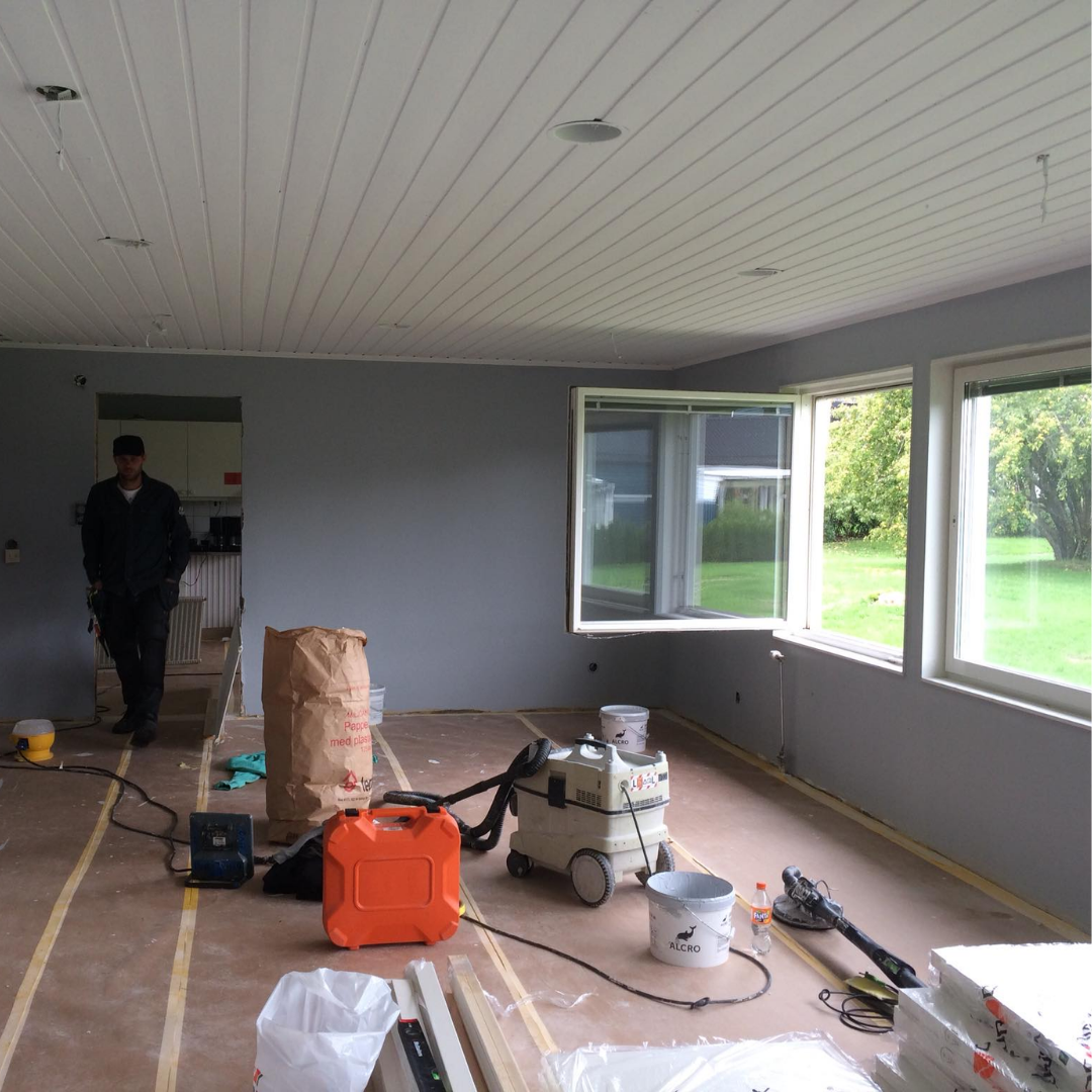 An interior room undergoing renovation, with gray painted walls, a white paneled ceiling, and exposed subflooring. Construction tools, including a vacuum, buckets, and an orange container, are scattered around. A man in dark clothing stands near a doorway in the background, suggesting a professional working on a home improvement project.