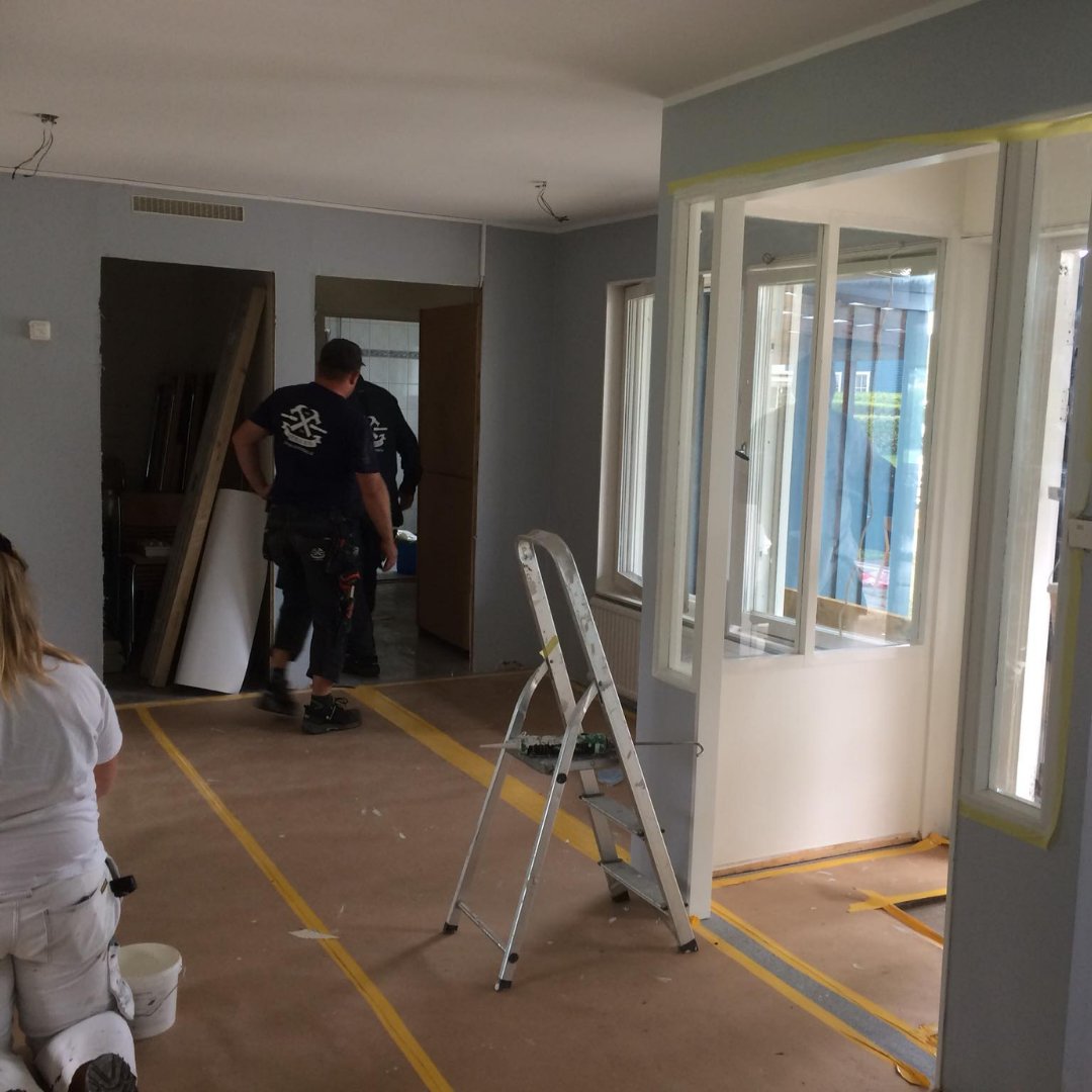 An interior room under renovation, with light gray walls and exposed subflooring marked with yellow tape. Two workers are visible in the background, and a stepladder stands in the foreground, indicating ongoing construction or remodeling work.