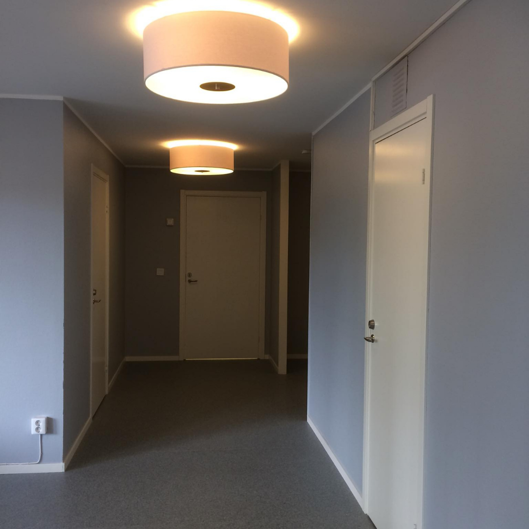 A brightly lit hallway with light gray walls and a gray floor, featuring multiple white doors leading to different rooms. Two modern, circular ceiling lights cast a warm glow, creating a clean and inviting interior space.