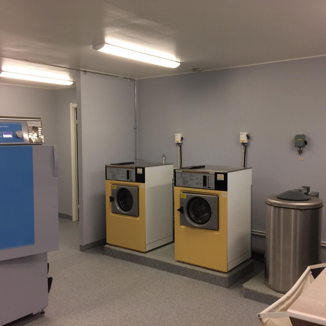 A clean and well-lit laundry room featuring two yellow and white commercial washing machines, a stainless steel bin, and other laundry equipment. The walls are light gray, and the floor is a gray speckled pattern, indicating a functional and organized communal laundry facility.
