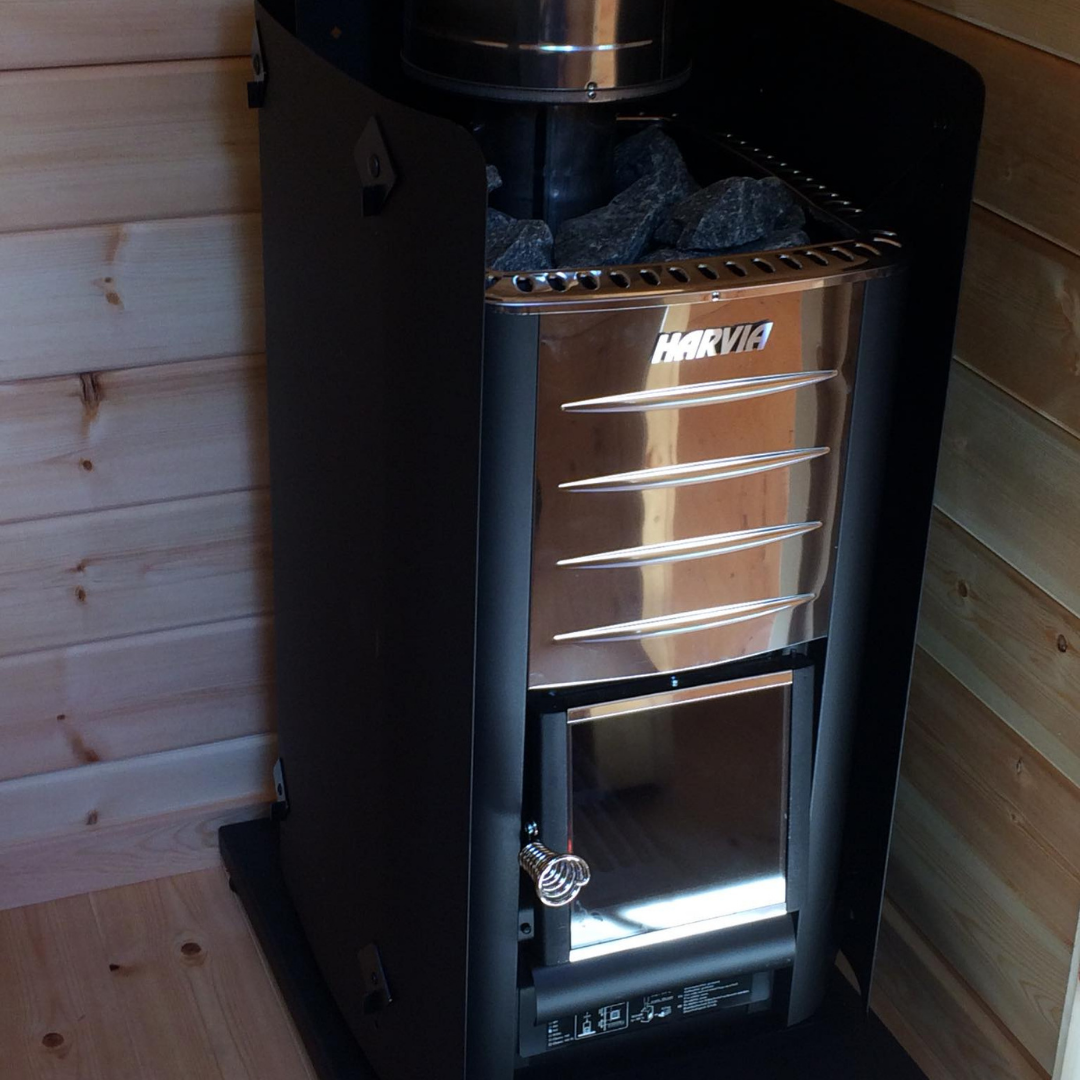 A modern black sauna heater with a stainless steel front panel and the brand name "HARVIA" visible. The heater is filled with dark gray sauna stones and is placed on a light wooden floor next to a wooden wall, highlighting a contemporary sauna interior.