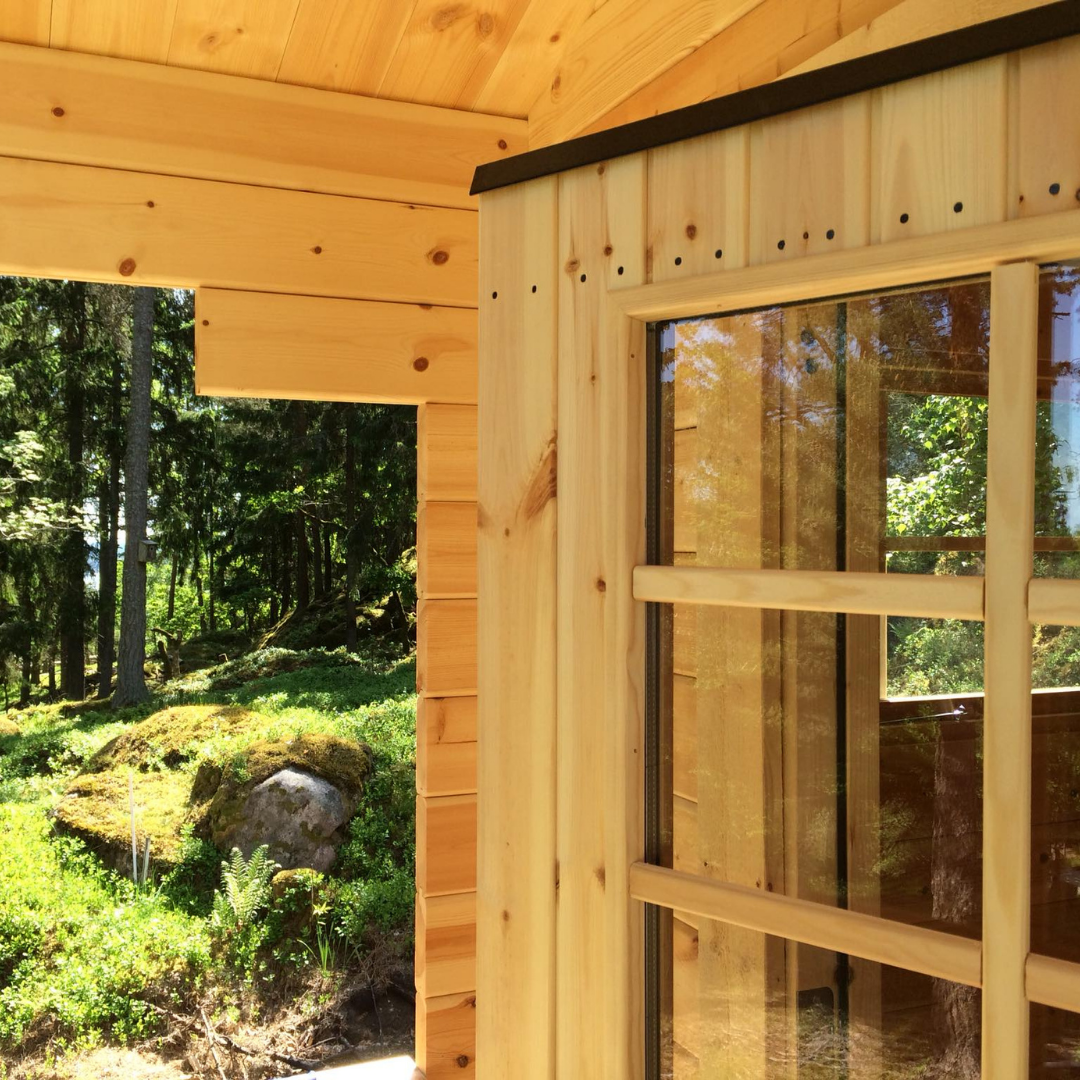 A close-up interior view of a wooden cabin, looking out through a window with wooden frames. The exterior shows a vibrant green forest with mossy rocks and tall trees, emphasizing the connection between the indoor space and nature.