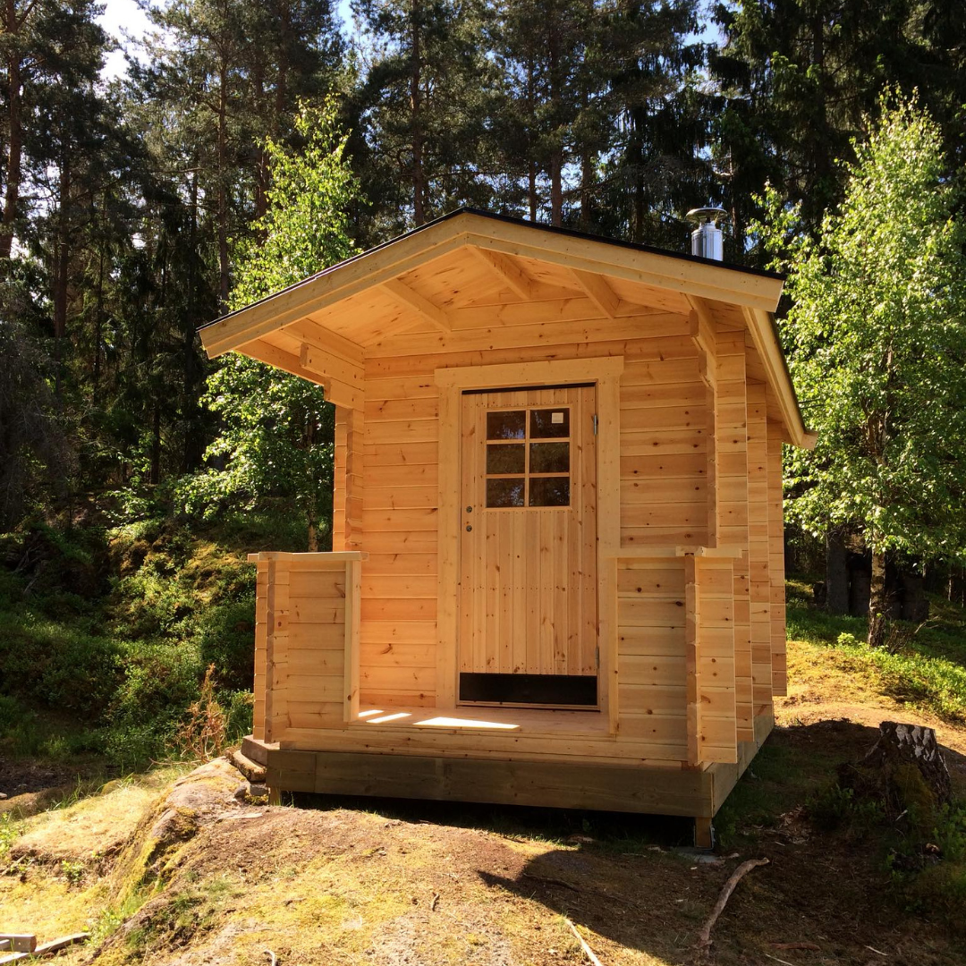 A newly built light wooden sauna cabin with a small covered porch and a wooden door with a window. The cabin is situated on a grassy, slightly elevated area surrounded by tall green trees under a bright sky, showcasing a rustic outdoor retreat.