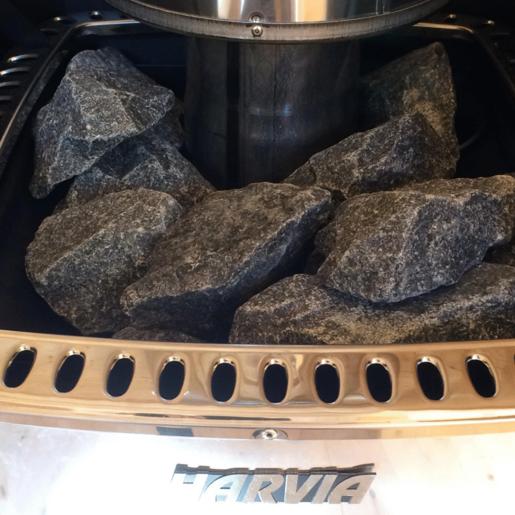 A close-up of a sauna heater filled with dark gray sauna stones, with the brand name "HARVIA" visible at the bottom. The metallic finish of the heater and the rough texture of the stones are prominent, indicating a high-quality sauna heating element.