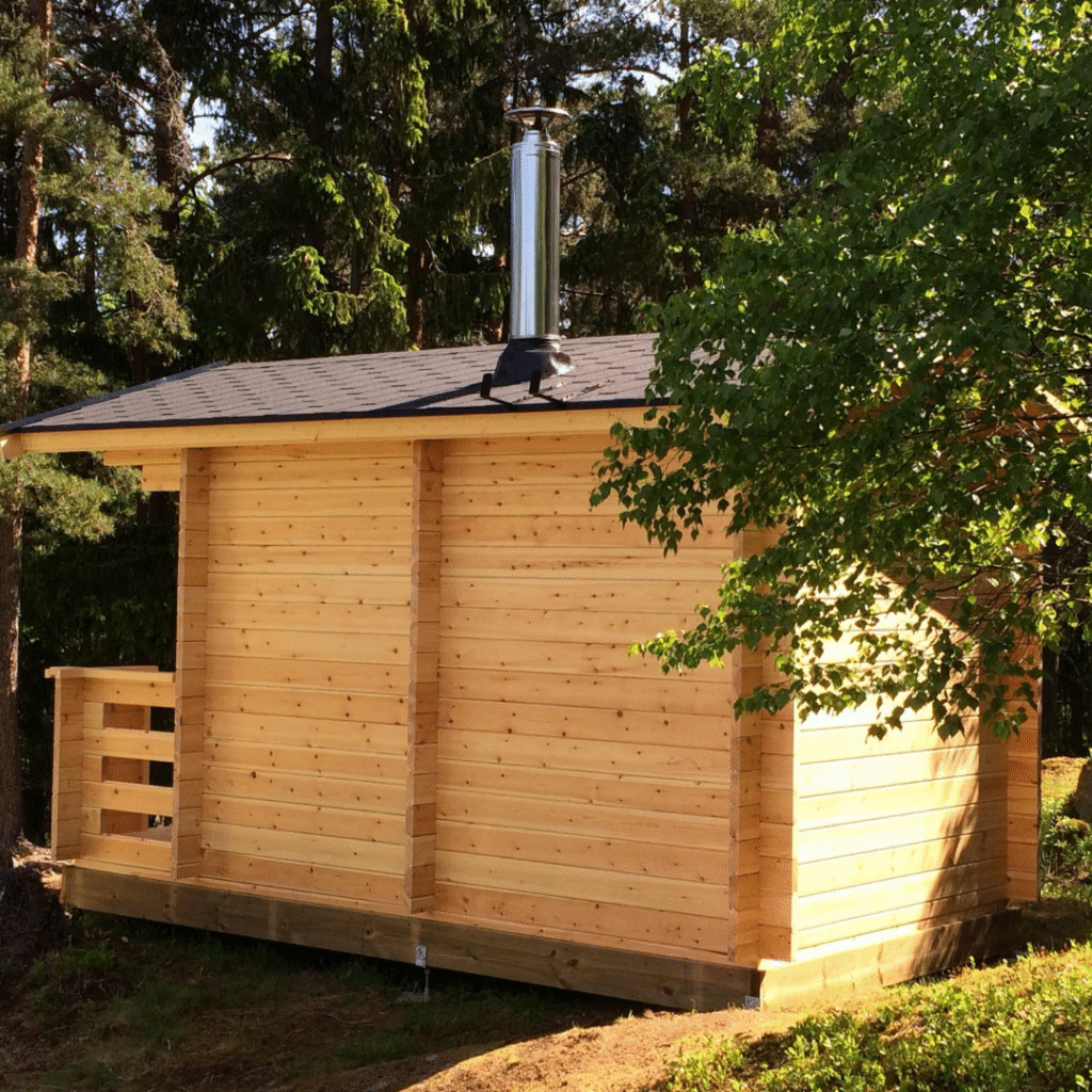 The side view of a newly built light wooden sauna cabin with a dark roof and a prominent metal chimney. The cabin is nestled among green trees, highlighting its natural setting and the simple, functional design of an outdoor sauna.