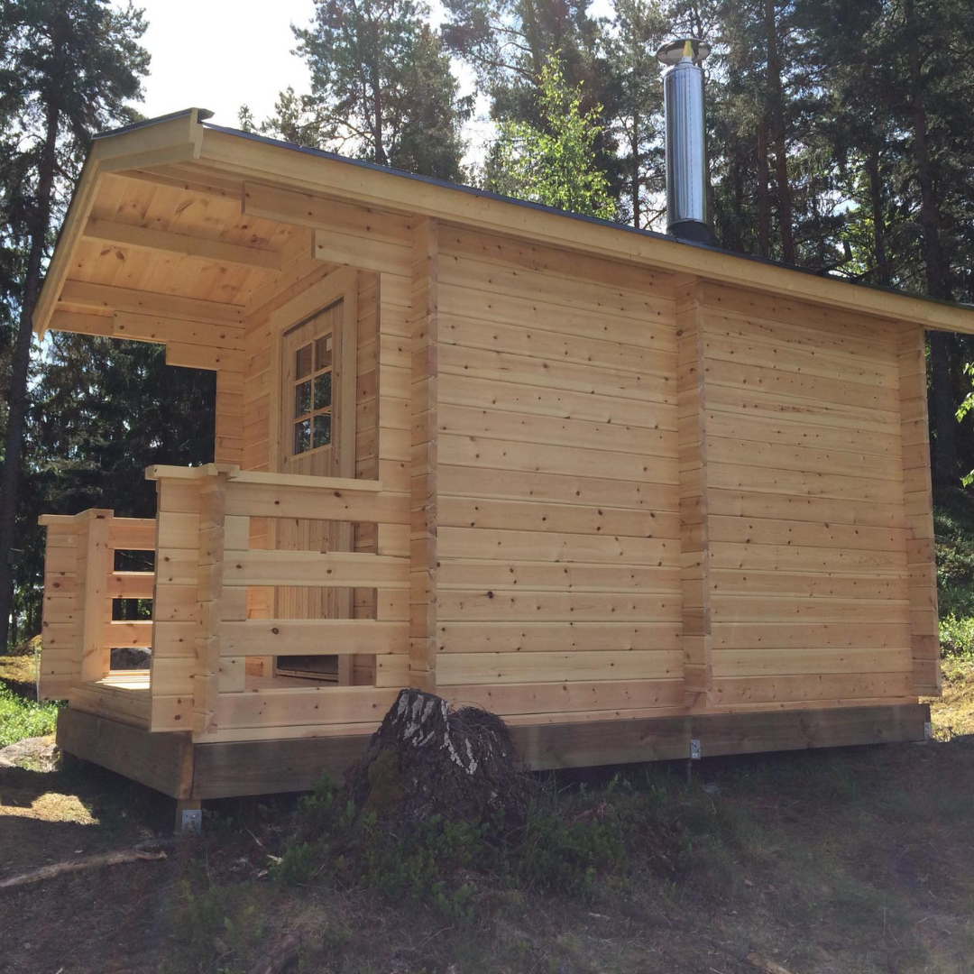 A newly constructed light wooden sauna cabin with a small porch and railing, featuring a wooden door with a window. A metal chimney extends from the roof, and the cabin is set on a raised foundation amidst green trees, showcasing a rustic outdoor sauna.