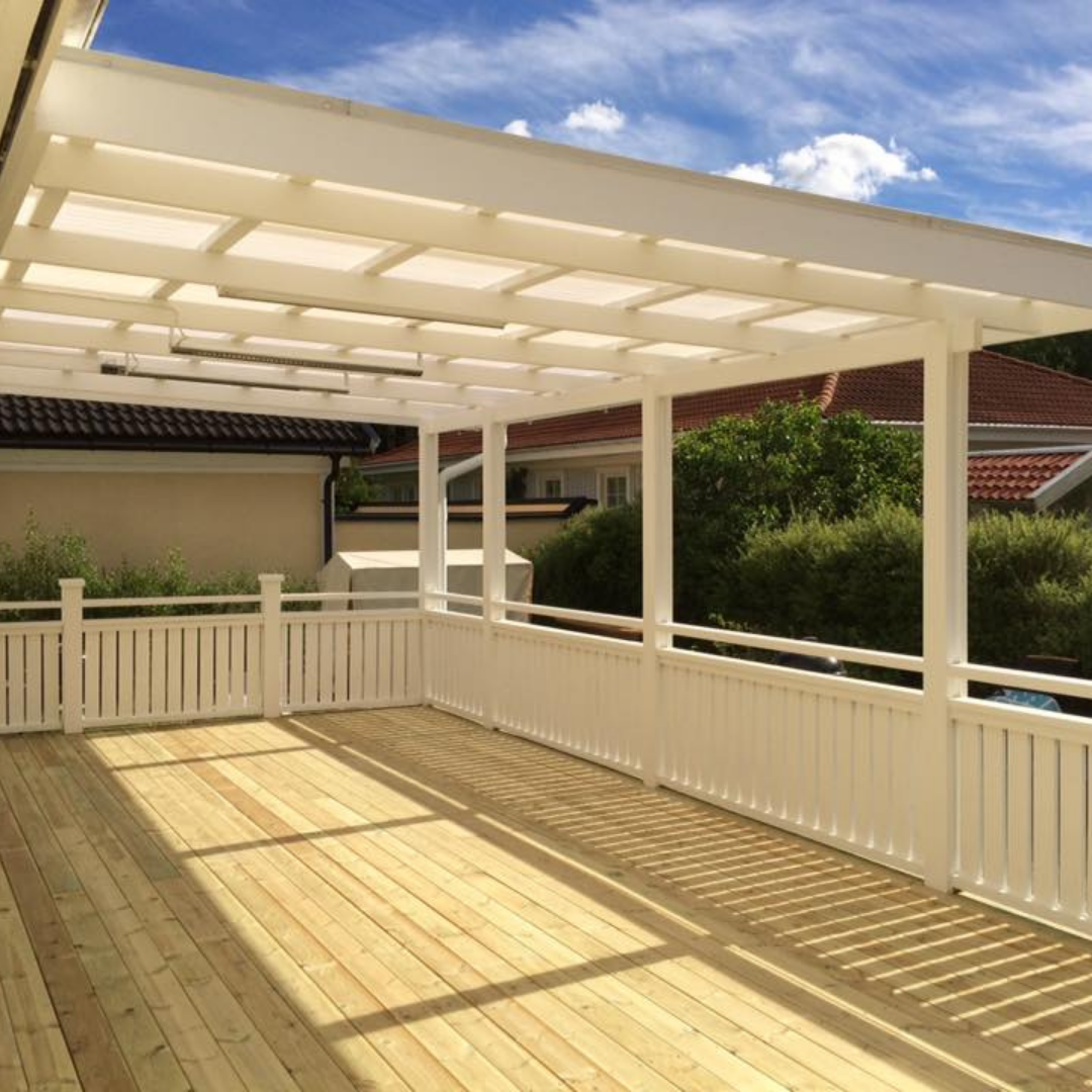 A spacious, newly constructed wooden deck with white railings and a matching white pergola overhead, providing shade. The deck features light-colored wooden planks and is set against a backdrop of residential houses and a clear blue sky, ideal for outdoor living spaces and home additions.