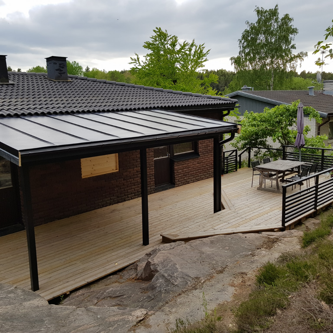 A large wooden deck with black railings extending from a brick house with a dark tiled roof. The deck features an outdoor dining set with an umbrella and is built on a rocky, uneven terrain, surrounded by green trees under an overcast sky, showcasing an expansive outdoor living area.
