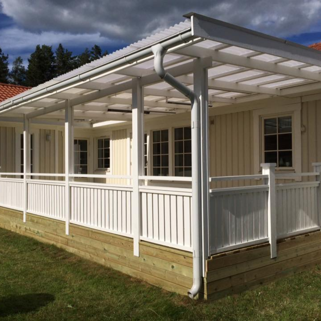 A light-colored wooden house with a spacious, newly built covered patio or veranda. The patio features a white wooden railing, white support pillars, and a translucent roof, providing an inviting outdoor living space next to a green lawn.